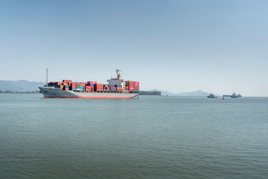 A large cargo ship travels across the open ocean, framed against distant mountains under a clear sky.
